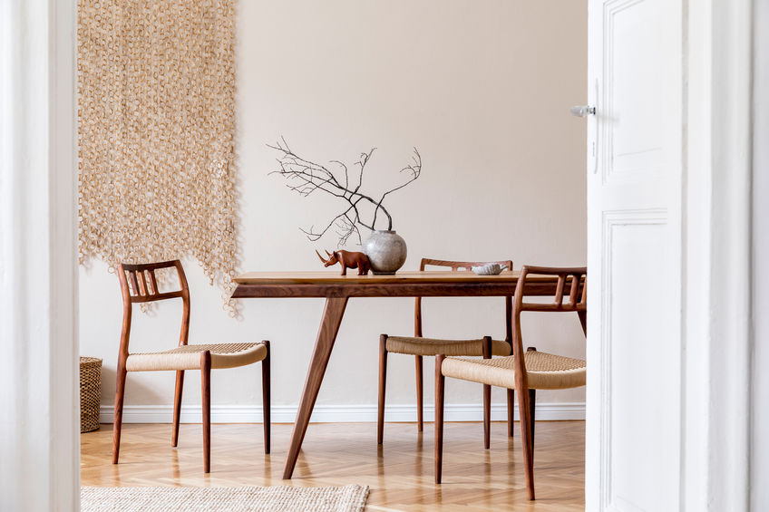Stylish and beige interior of dining room with design wooden table and chairs, vase with flowers, sculpture, elegant and rattan accessories.